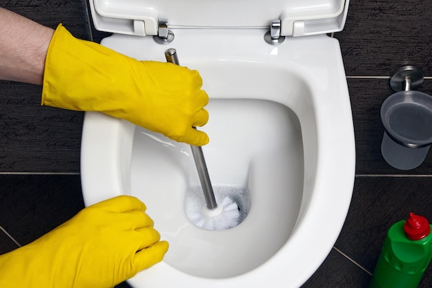 Mężczyzna w żółtych gumowych rękawiczkach czyści toaletę pędzlem i środkiem dezynfekującym