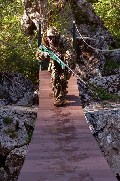 Zdjęcie mężczyzna wojskowy lub gracz airsoft w garniturze kamuflażowym skrada się przez most linowy i celuje z karabinu snajperskiego na bok lub do celu