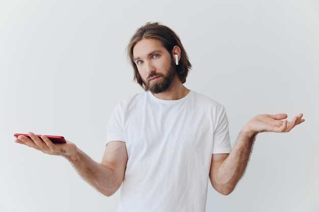 Mężczyzna z blogerem z brodą w białej koszulce z telefonem i słuchawkami bezprzewodowymi patrzy w telefon i rozkłada ręce na białym tle na białym tle