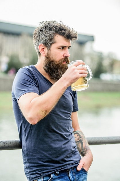 Mężczyzna z brodą i wąsami trzyma szklankę piwa na zewnątrz Facet odpoczywa przy zimnym piwie z beczki Hipster zrelaksowany pije piwo na świeżym powietrzu Kawiarnia letni taras Jasne ale lub ciemne stouty piją wszystko