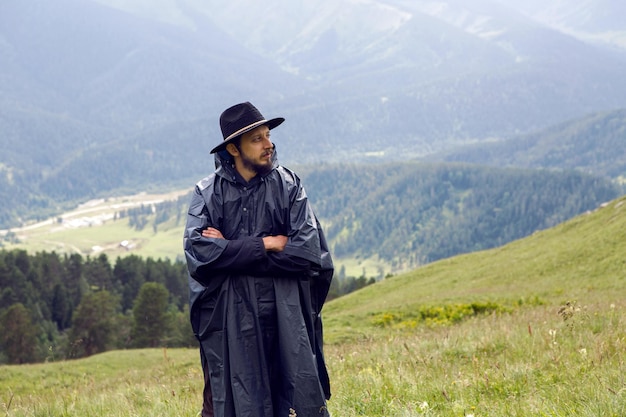 Zdjęcie mężczyzna z brodą pasterza stojący w górach w czarnym płaszczu przeciwdeszczowym w deszczu