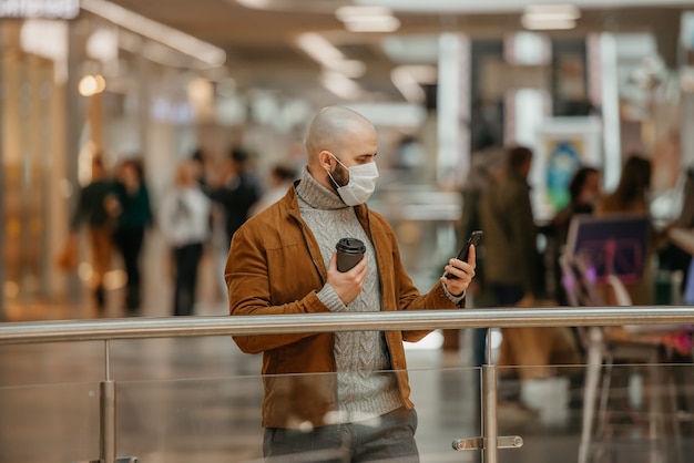 Mężczyzna z brodą w masce na twarz, aby uniknąć rozprzestrzeniania się koronawirusa, używa smartfona i trzyma filiżankę kawy w centrum handlowym. Łysy facet w masce chirurgicznej zachowuje dystans społeczny.