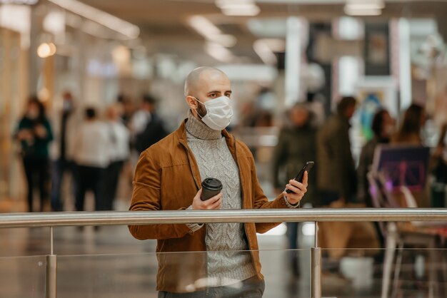 Mężczyzna z brodą w masce na twarz patrzy w lewo i trzyma smartfon i filiżankę kawy w centrum handlowym. Łysy facet w masce chirurgicznej zachowuje dystans społeczny.