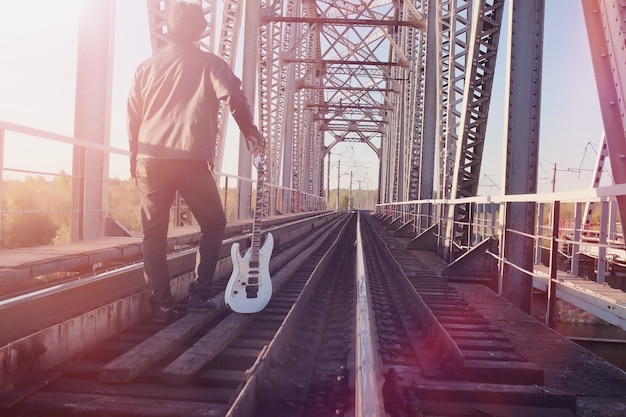 Mężczyzna z gitarą elektryczną na kolei. Muzyk w skórzanej kurtce z gitarą na ulicy w strefie przemysłowej. Gitarzysta na moście.