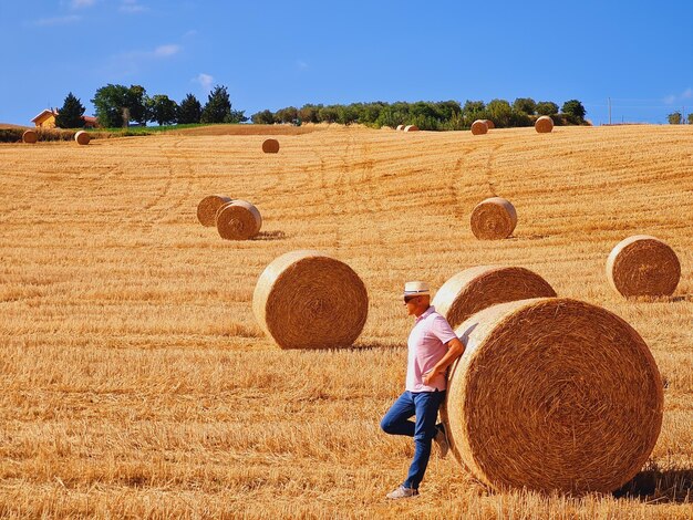 Zdjęcie mężczyzna z kapeluszem opierający się o balę siana na zbiorowym polu pszenicy