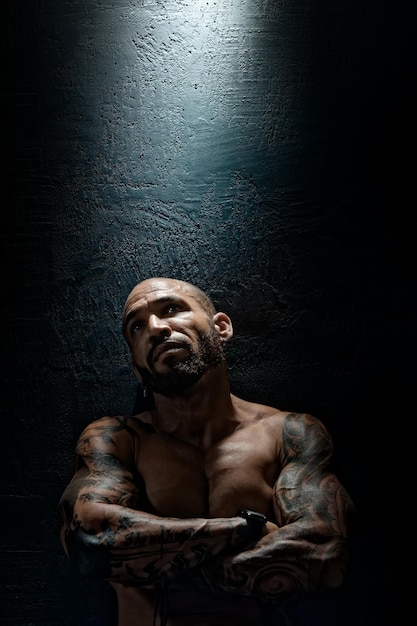 Mężczyzna z nagim ciałem w tatuażach stoi pod ścianą i krzyżuje ręce na piersi Concept