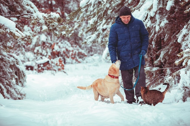 Mężczyzna z psem na smyczy i kotem spaceruje zimą po zaśnieżonym sosnowym lesie