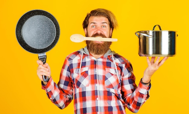 Zdjęcie mężczyzna z rondelkiem i patelnią w rękach brodaty szef kuchni z drewnianą łyżką w naczyniu do gotowania w ustach