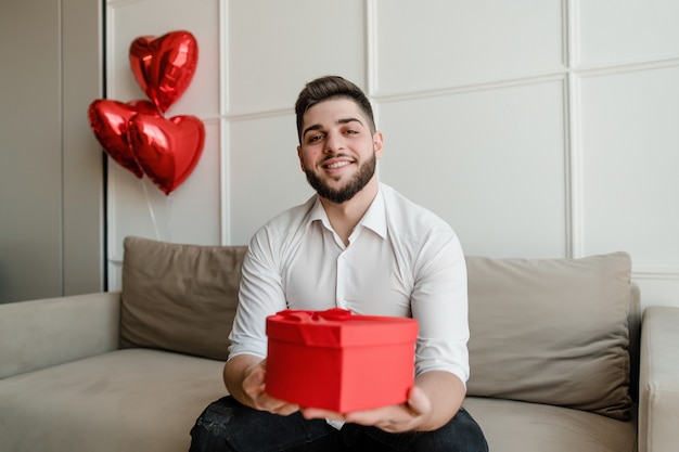 Mężczyzna z sercem w kształcie czerwieni teraźniejszości w prezenta pudełka obsiadaniu na leżance z balonami w domu