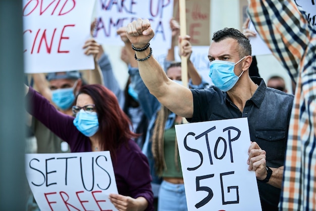 Mężczyzna z uniesioną pięścią w masce ochronnej i trzymający transparent z napisem „stop 5G” na publicznych demonstracjach