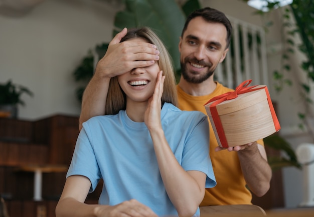 Mężczyzna zakrywający oczy swojej dziewczyny, dając prezent pudełko. Urocza para siedzi razem w kawiarni, randka romańska