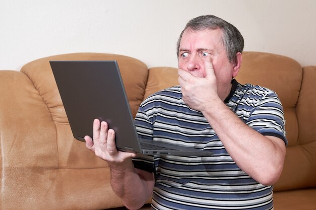 Mężczyzna ze zdziwioną twarzą trzyma w rękach laptopa siedząc na kanapie