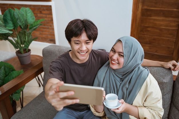 Mężczyźni I Dziewczęta W Hidżabie Patrzą Na Ekran Smartfona, Robiąc Selfie, Uśmiechając Się I Trzymając Kubki W Salonie