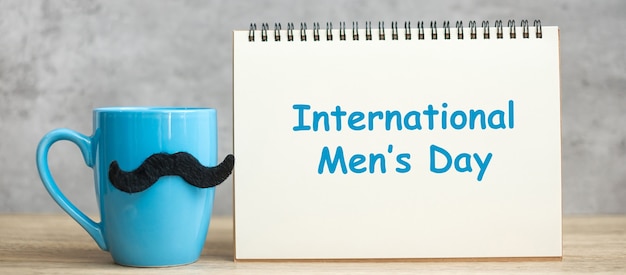 Zdjęcie międzynarodowy dzień mężczyzn z papierowym notatnikiem, niebieska filiżanka kawy lub kubek herbaty i wystrój czarne wąsy na stole. szczęśliwy dzień ojca i koncepcja uroczystości