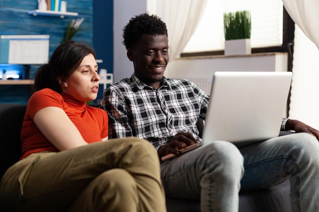 Międzyrasowy zrelaksowany para patrząc na ekran laptopa siedząc na kanapie w salonie. Młodzi ludzie korzystający z internetu i mediów społecznościowych, korzystający z komputera podczas oglądania śmiesznych filmów
