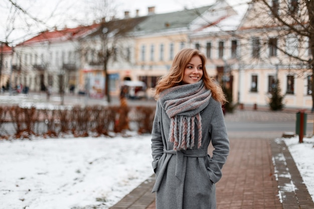 Miejska ładna młoda kobieta z pięknym uśmiechem w szarym stylowym płaszczu w ciepłym szaliku z dzianiny spaceruje po mieście w pobliżu zabytkowych budynków w zimowy dzień