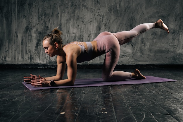Mięśni młoda kobieta lekkoatletycznego z doskonałe piękne ciało noszenia odzieży sportowej ćwiczenia pośladki nogi na macie do jogi. Kobieta fitness kaukaski pozowanie Studio z ciemnoszarym tle.