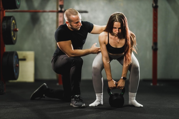 Mięśni młoda kobieta pracuje z osobistym trenerem na siłowni. Trener pomaga kobiecie w treningu z Kettlebell.