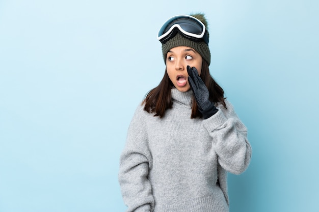 Mieszana narciarz wyścigu dziewczyna w okularach snowboardowych nad izolowanym niebieskim szeptem coś z gestem zaskoczenia, patrząc w bok.