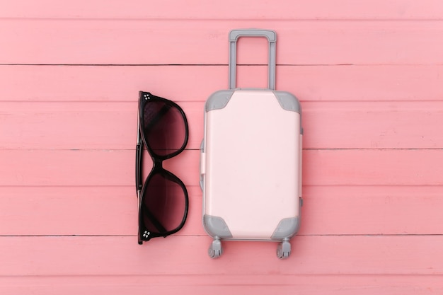 Mieszkanie świeckich wakacje wakacje i koncepcja planowania podróży. Mini walizka podróżna z tworzywa sztucznego i okulary przeciwsłoneczne na różowym tle drewnianych. Widok z góry