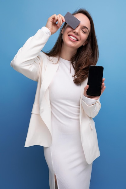 Miła miła brunetka młoda kobieta w sukience i kurtce z plastikową kartą płatniczą ze smartfonem
