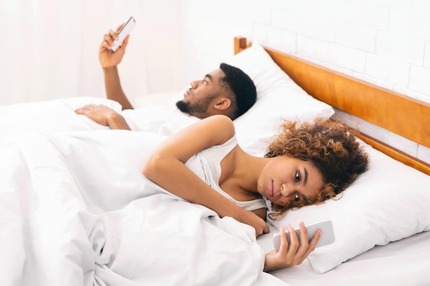 Milenijna para leżąca na łóżku plecami do pleców ze smartfonami