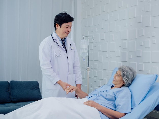 Miły azjatycki lekarz mężczyzna w białym garniturze odwiedza rozmowy i udziela wsparcia, trzymając rękę szczęśliwej starszej kobiety starszej kobiety w jasnoniebieskiej sukience leżącej na łóżku w roztworze soli fizjologicznej w sali szpitalnej