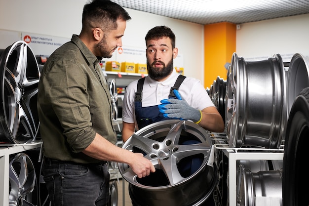 Zdjęcie miły klient i pewny siebie mechanik samochodowy opowiadający o kołach samochodowych, w sklepie, stojący wśród stojaków na felgi