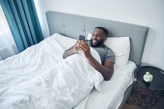 Miły międzynarodowy mężczyzna odczuwający szczęście podczas czytania dobrych wiadomości w Internecie, leżąc na łóżku