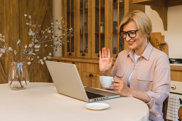 Miły poranek. Wesoła uśmiechnięta starsza kobieta w wieku stojąca w kuchni i korzystająca z laptopa podczas odpoczynku po śniadaniu, freelancer pracujący w domu.