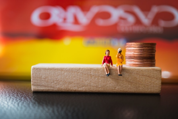 Miniaturowi ludzie, pary kobiety obsiadanie na drewnianym bloku z stert monetami używać jako biznes i financ