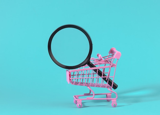 Miniaturowy metalowy różowy wózek i czarna plastikowa lupa na jasnoniebieskiej powierzchni. Pojęcie wyszukiwania i selekcji zakupów, oszczędności