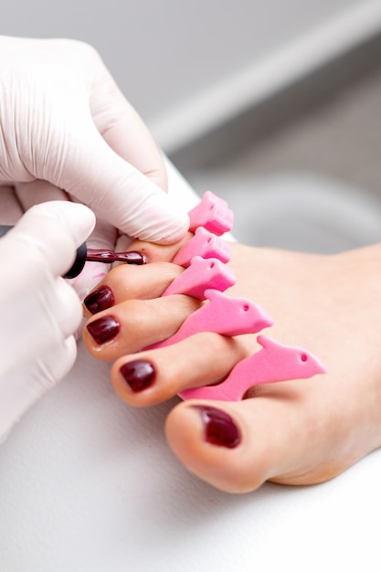 Mistrz manicure maluje na kobiecych paznokciach bordowym lakierem do paznokci pędzlem w białych rękawiczkach