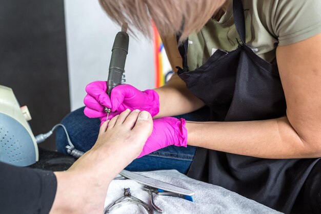 Mistrz Pedicure W Różowych Rękawiczkach Nacina Naskórek I Paznokcie Szelakowe W Salonie Pedicure Wiertarką Profesjonalny Pedicure W Gabinecie Kosmetologicznym Higiena Stóp W Gabinecie Kosmetycznym