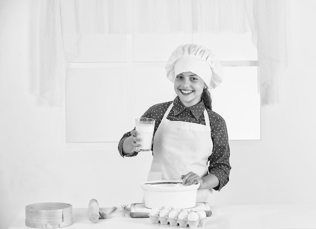 Mleko jako składnik dziecko w mundurze szefa kuchni i kapeluszu nastoletnia dziewczyna przygotowuje ciasto robiąc ciasto według przepisu czas jeść szczęśliwe dziecko gotowanie w kuchni piec ciasteczka w kuchni profesjonalny i wykwalifikowany piekarz