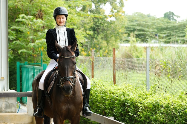 Młoda 20s Azjatycka kobieta w garniturze jeździeckim z pięknym zwierzęciem na zewnątrz. Kobieta jeździ na wysokim, inteligentnym, silnym koniu w stajni i trenuje naukę na zielonym tle na całej długości