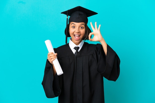Młoda absolwentka uniwersytetu dziewczyna na odosobnionym niebieskim tle pokazując znak ok i gest kciuka w górę