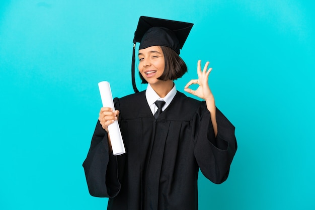 Młoda absolwentka uniwersytetu dziewczyna nad odosobnioną niebieską ścianą pokazującą znak ok palcami