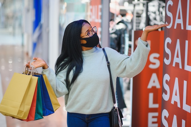 Młoda African American kobieta w centrum handlowym w nowej normalności po covid-19 - szczęśliwa i piękna czarna dziewczyna w masce na twarz, trzymając torby na zakupy, ciesząc się w sklepie z modą urody