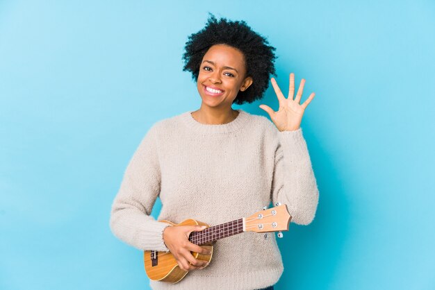 Młoda Afroamerykanin kobieta gra ukelele na białym tle uśmiechnięty wesoły pokazując numer pięć palcami.
