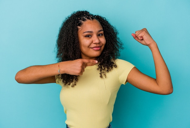 Młoda Afroamerykanin kobieta na białym tle na niebieskiej ścianie pokazuje gest siły z rękami, symbol kobiecej mocy