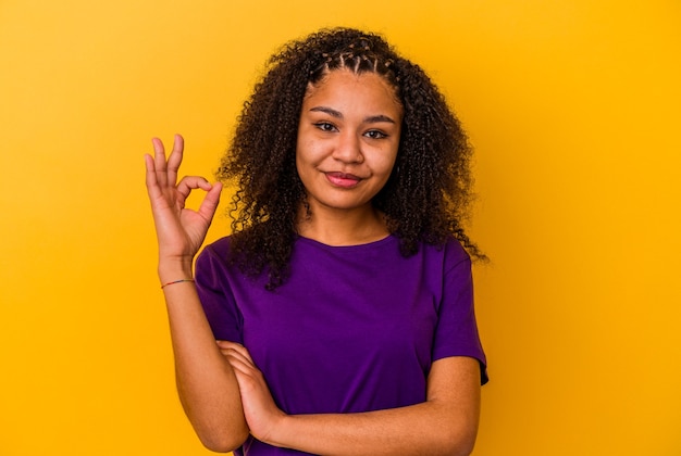 Młoda Afroamerykanin kobieta na żółtej ścianie mruga okiem i trzyma w porządku gest ręką