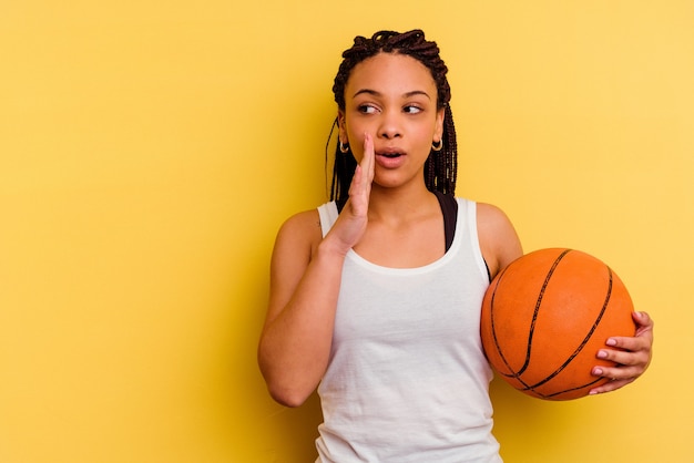 Młoda Afroamerykanka gra w koszykówkę na żółtym tle mówi tajną gorącą wiadomość o hamowaniu i patrzy na bok