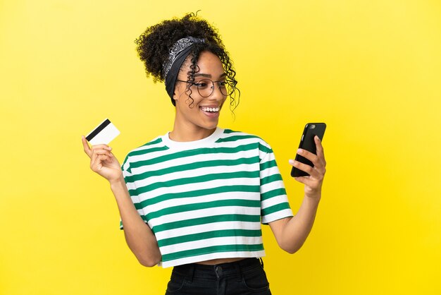 Młoda afroamerykanka na białym tle na żółtym tle kupując za pomocą telefonu komórkowego za pomocą karty kredytowej