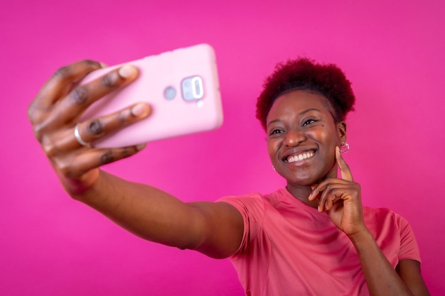 Młoda Afroamerykanka odizolowana na różowym tle uśmiecha się i robi selfie w studio