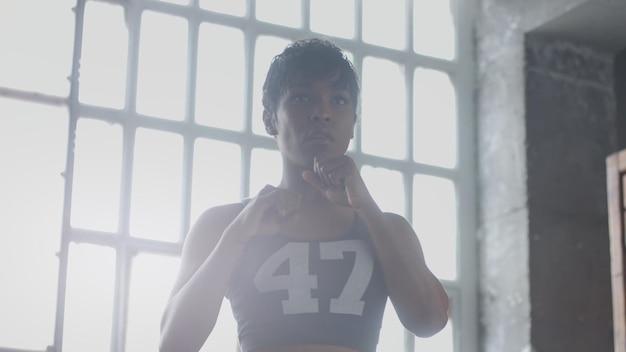 Młoda afroamerykanka rasy mieszanej robi trening bokserski w słonecznym lofcie Konturowe światło patrząc przez kamerę