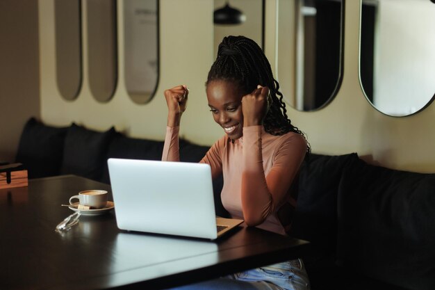 Zdjęcie młoda afroamerykanka siedzi przy stole w kawiarni w pobliżu laptopa, podnosząc pięści, wyrażając sukces wi