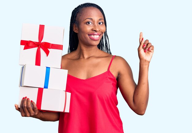 Młoda Afroamerykanka trzyma prezent zaskoczona pomysłem lub pytaniem wskazującym palcem z radosną twarzą, numer jeden
