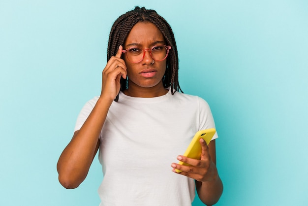 Młoda Afroamerykanka trzyma telefon komórkowy na białym tle na niebieskim tle, wskazując palcem świątynię, myśląc, skupioną na zadaniu.