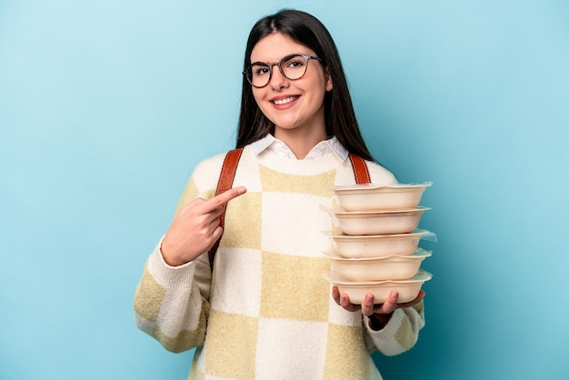 Młoda Afroamerykanka trzyma Tupperware na białym tle na niebieskim tle, uśmiechając się i wskazując na bok, pokazując coś w pustej przestrzeni